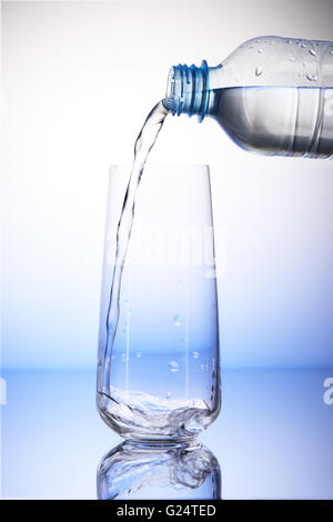 Water pouring from plastic bottle into empty drinking glass with reflection on white and blue background Stock Photo