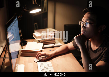 Pensive asian young man in glasses working with computer and thinking in the evening at home Stock Photo