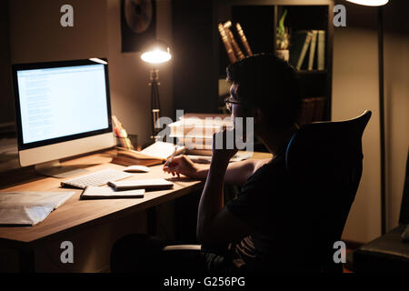Thoughtful asian young man sitting and studying using computer in dark room at home Stock Photo
