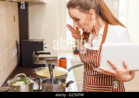 Young woman trying cooking with recipe from laptop. Shocked caucasian woman online learning cooking Stock Photo