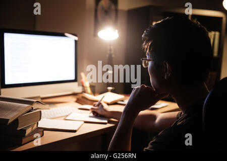 Focused asian young man in glasses sitting and using computer for studying in dark room Stock Photo