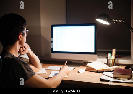 Back view of serious asian young man reading information from blank screen computer and making notes in the evening Stock Photo