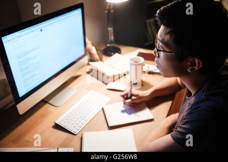 Thoughtful asian young man reading and writing using information from computer in dark room Stock Photo