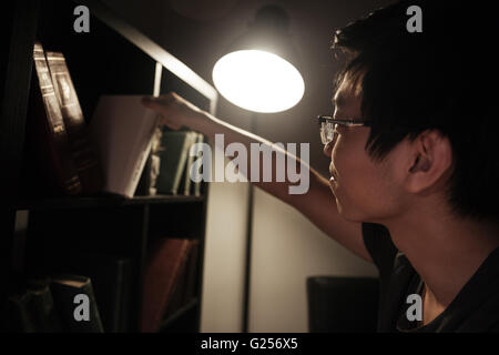 Handsome asian young man in glasses choosing book from bookshelf for reading in the evening at home Stock Photo