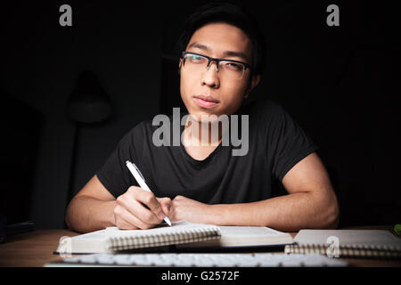 Bored serious asian young man in glasses learning and writing in the evening at home Stock Photo
