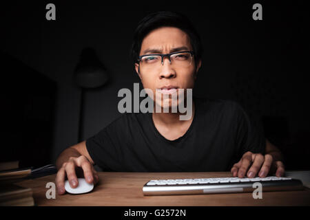 Concentrated asian young man in glasses working with computer in dark room Stock Photo