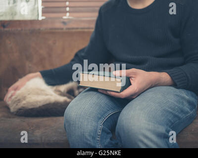 A young man is sitting on a sofa with a sleeping cat and is holding a big green book Stock Photo