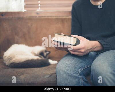A young man is sitting on a sofa with a sleeping cat and is holding a big green book Stock Photo