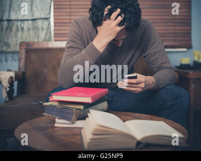 A young man surrounded by books is sitting on a sofa and is looking upset as he is using his smartphone Stock Photo