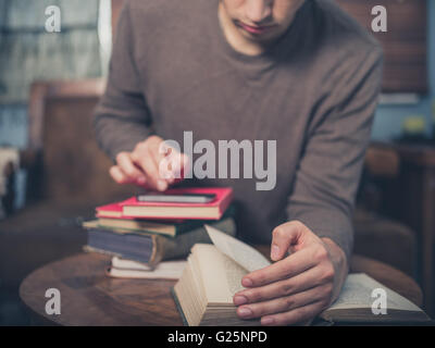 A young man is sitting on a sofa surrounded by books and is using his smartphone Stock Photo