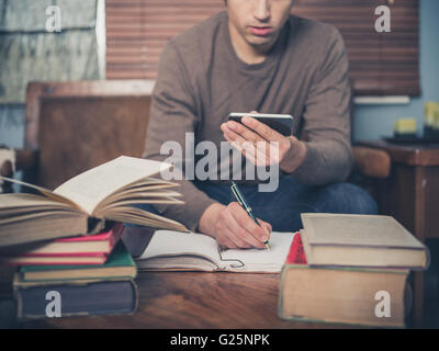 A young man is sitting on a sofa surrounded by books and is using his smartphone Stock Photo