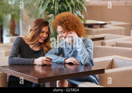 Two happy lovely young women using thir smartphones in cafe and laughing Stock Photo