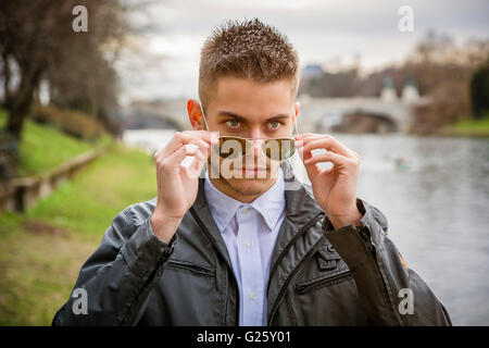 Three-quarter length of contemplative light brown haired young man wearing grey jacket and denim jeans standing beside picturesq Stock Photo