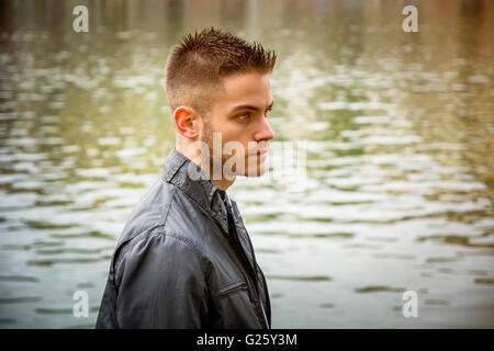 Three-quarter length of contemplative light brown haired young man wearing grey jacket and denim jeans standing beside picturesq Stock Photo