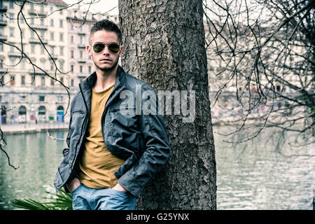 Three-quarter length of contemplative light brown haired young man wearing grey jacket and denim jeans standing beside picturesq Stock Photo
