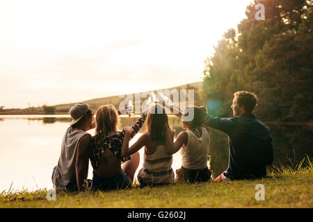 Group of young people sitting in a row at a lake with beers. Young friends toasting and celebrating with beers at the lake on a Stock Photo