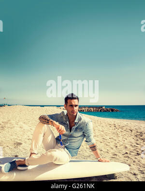 Stylish handsome young man in mirrored sunglasses sitting on surfboard on beach in sunlight Stock Photo