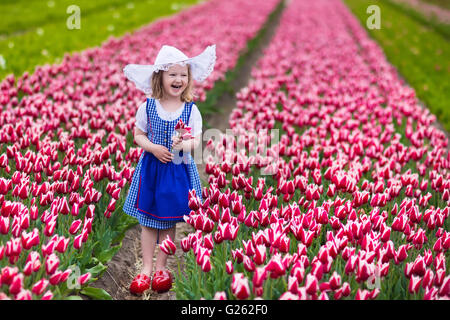 Adorable little girl wearing Dutch traditional national costume dress, wooden clogs and hat playing in a field of blooming tulip Stock Photo