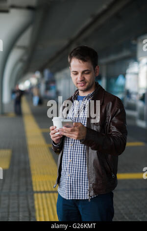 Man Using Mobile Phone near the airport terminal with a cup of coffee waiting for the taxi Stock Photo