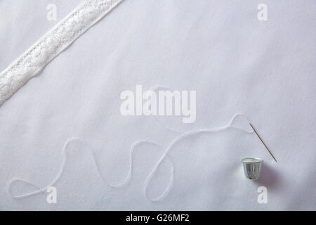Background sewing needle threaded with white thread thimble and lace ribbon on white cloth. Horizontal composition. Top view. Stock Photo