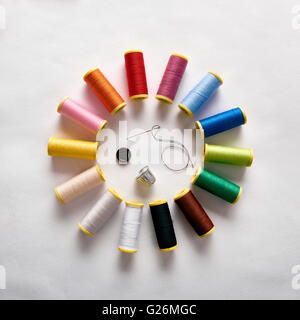 Spools of thread in circle, button, needle and thimble on white cloth. Square composition. Top view Stock Photo