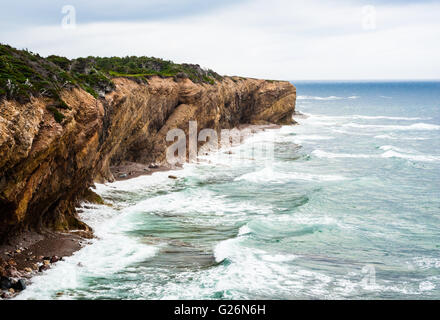 Waves splashing against eroding muddy coastline showing diagonal strata layers, under overcast sky, in Newfoundland, Canada. Stock Photo