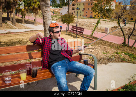 Guy documenting his hangover on a park bench with a selfie, a concept Stock Photo