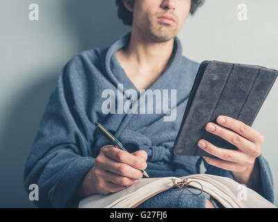 A young man wearing a bathrobe is using a tablet computer and is taking notes Stock Photo