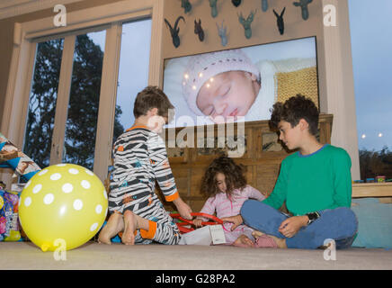 Birthday morning two year old in pajamas in lounge looking at gifts with siblings balloon and image of little girl as baby on tv Stock Photo