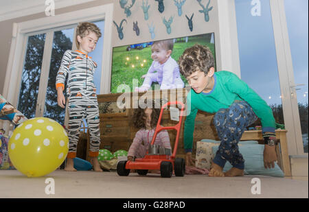 Birthday morning two year old in pajamas in lounge looking at gifts with siblings balloon and image of little girl as baby on tv Stock Photo