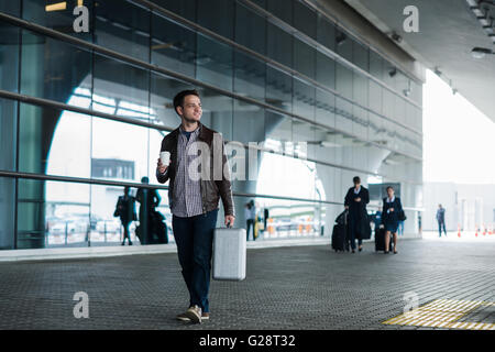 Portrait of a young man walking near the airport with suitcase and coffee Stock Photo