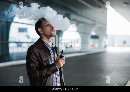 Young man enjoying a satisfying e-cigarette standing in profile against airport terminal background Stock Photo