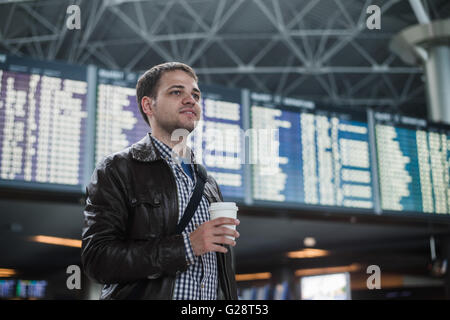 Young man with a bag in airport near flight timetable holding cup of coffee Stock Photo