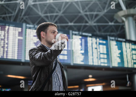 Young man with a bag in airport near flight timetable drinking coffee Stock Photo