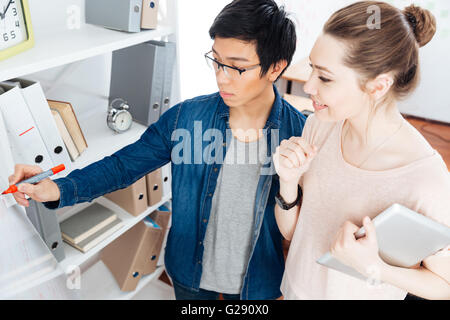 Smiling young businesswoman with tablet and focused asian man working and writing on flipchart in office Stock Photo