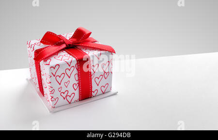 Decorative gift box with red ribbon and hearts printed on white table. Top view left. Grey background. Horizontal composition. Stock Photo
