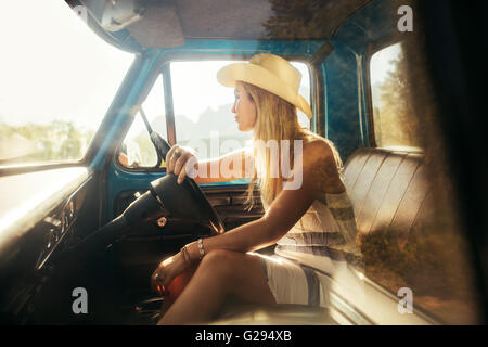 Portrait of young woman sitting on driving seat of a car and looking away. Girl wearing hat on a road trip. Stock Photo
