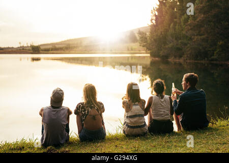 Rear view image of young people sitting in a row by a lake and looking at sunset. Group of friends sitting by a lake and relaxin Stock Photo