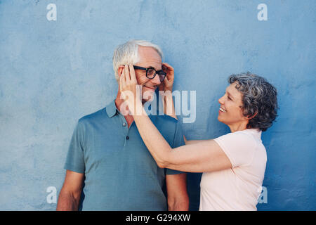 Portrait of woman adjusting the eyeglasses on her husband against blue background. Mature couple together on a blue wall. Stock Photo