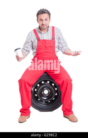 Confused auto mechanic holding coffee cup while sitting on car wheel taking a break Stock Photo