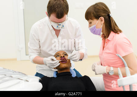 Male dentist with his assistant examining female patient's mouth sitting on chairs in clinic. Stock Photo