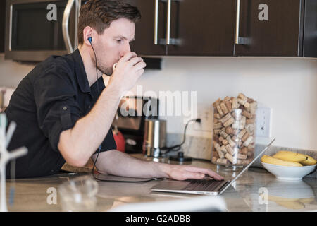 Man drinking while working on laptop computer in kitchen Stock Photo