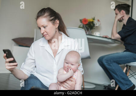 Mother holding baby (2-5 months) while using smart phone and father working in background Stock Photo