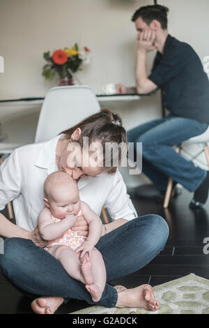 Mother holding baby (2-5 months) and father working in background Stock Photo