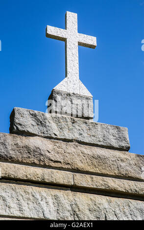 Smooth light gray almost white granite cross on top of rough hewn earth tone stone brick structure against clear cloudless blue sky Stock Photo