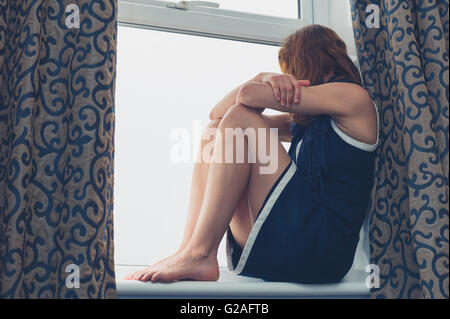 A young woman is sitting on a window sill and is looking out on a rainy day Stock Photo