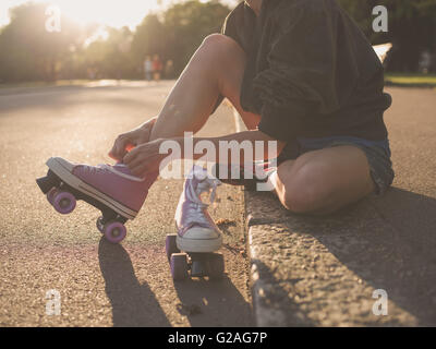 A young woman is sitting on the ground and is putting on roller skates in the park at sunset Stock Photo