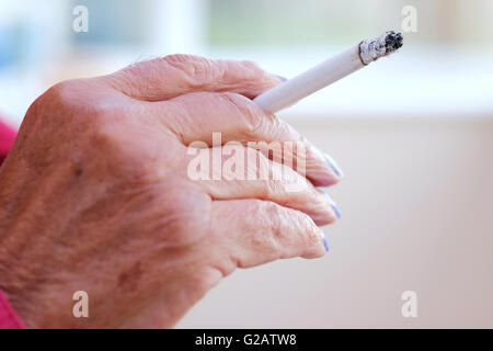 A close up of  An elderly female hand holding a lighted cigarette as it burns. There is an ash covered tip as the cigarette slowly burns Stock Photo