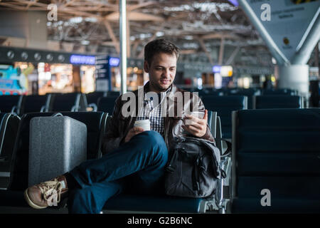 Handsome smiling man in casual wear holding luggage and messaging through his mobile phone while sitting at the hall of airport with coffee cup Stock Photo