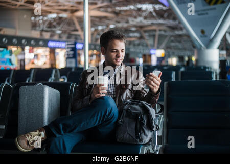 Handsome smiling man in casual wear holding luggage and messaging through his mobile phone while sitting at the hall of airport with coffee cup Stock Photo